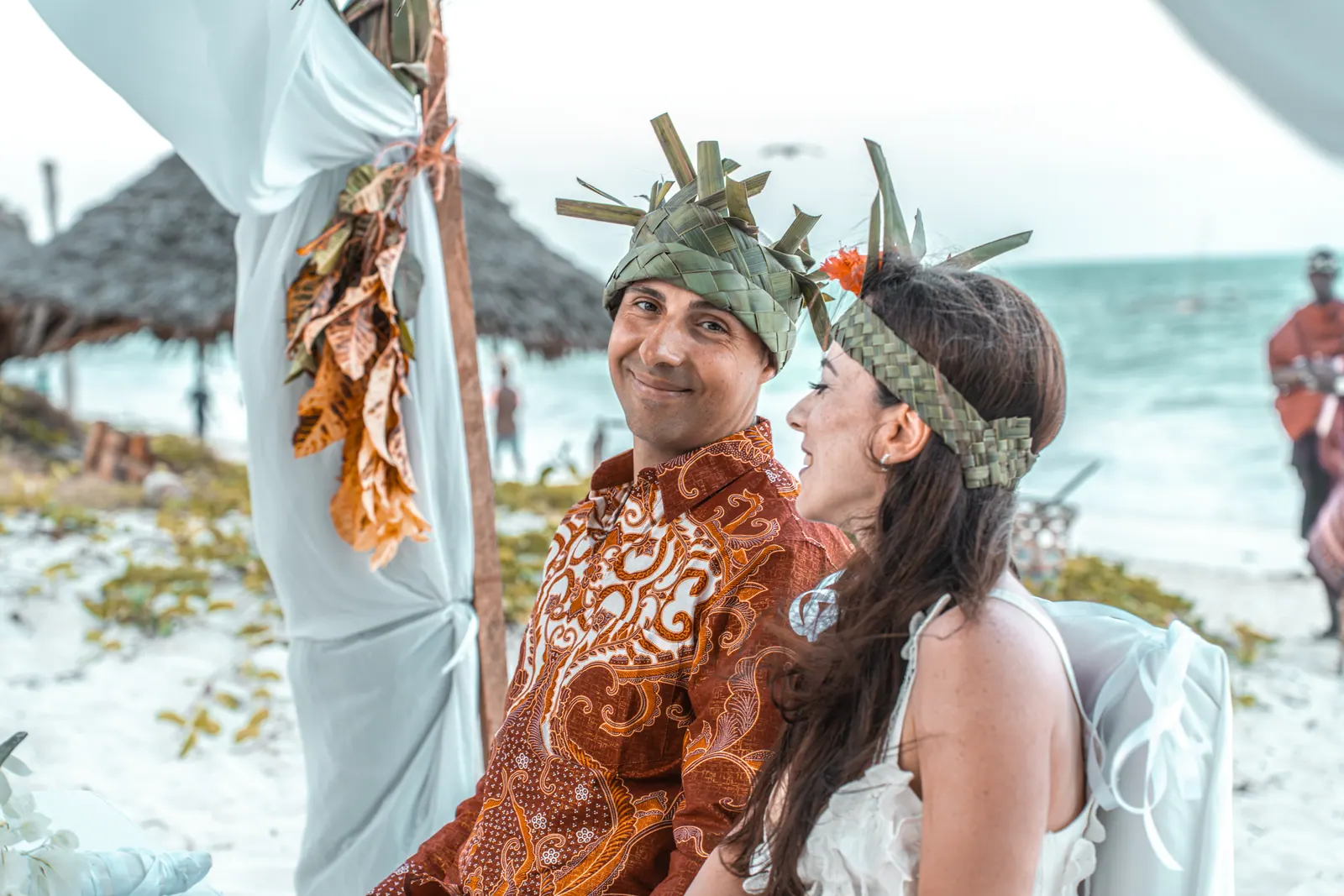 Bride and groom in palm-leaf crowns smiling at each other at a beach ceremony